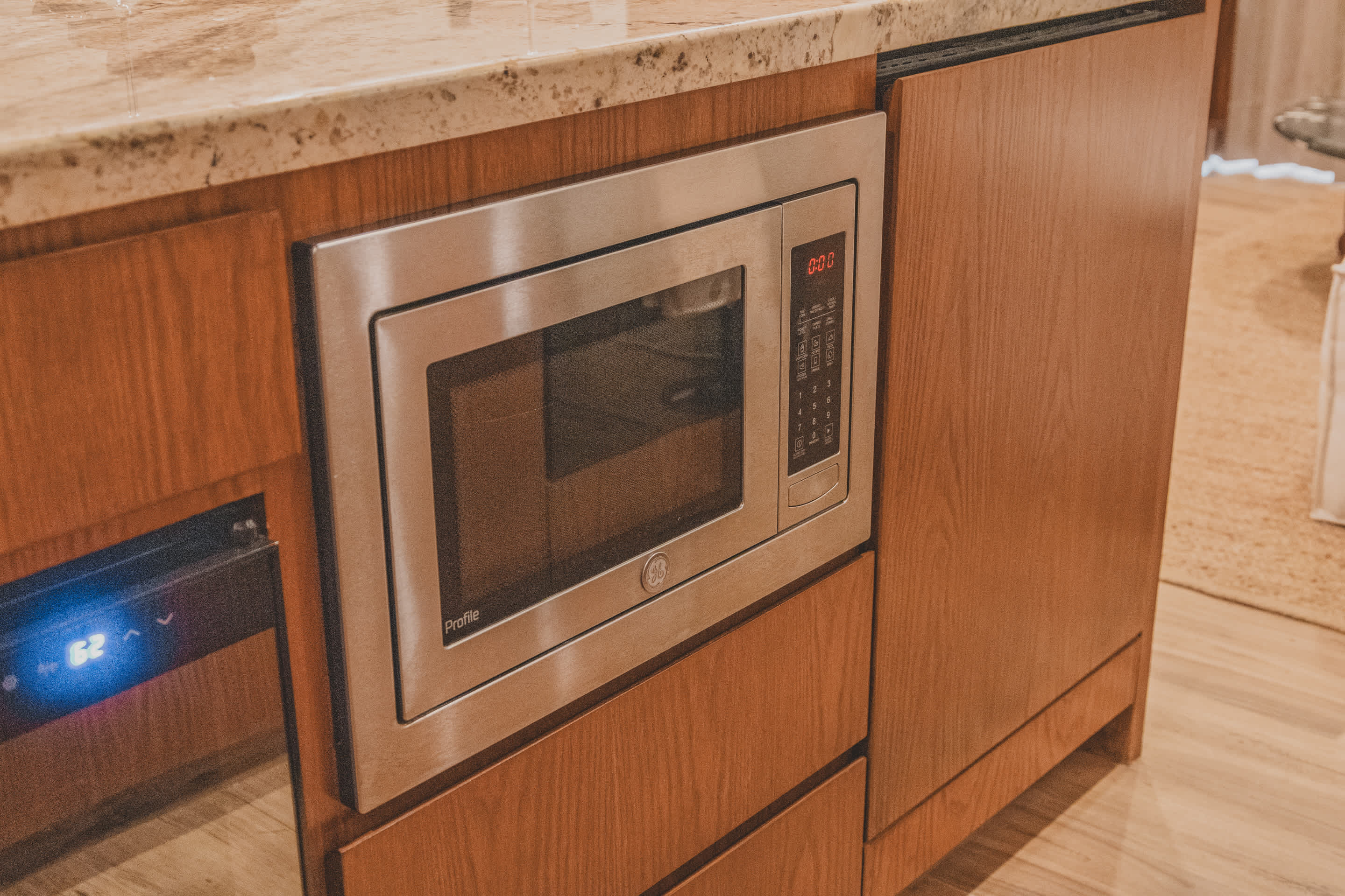 A close-up of a built-in stainless steel GE Profile microwave oven integrated into wooden kitchen cabinetry, set below a light-colored granite countertop.