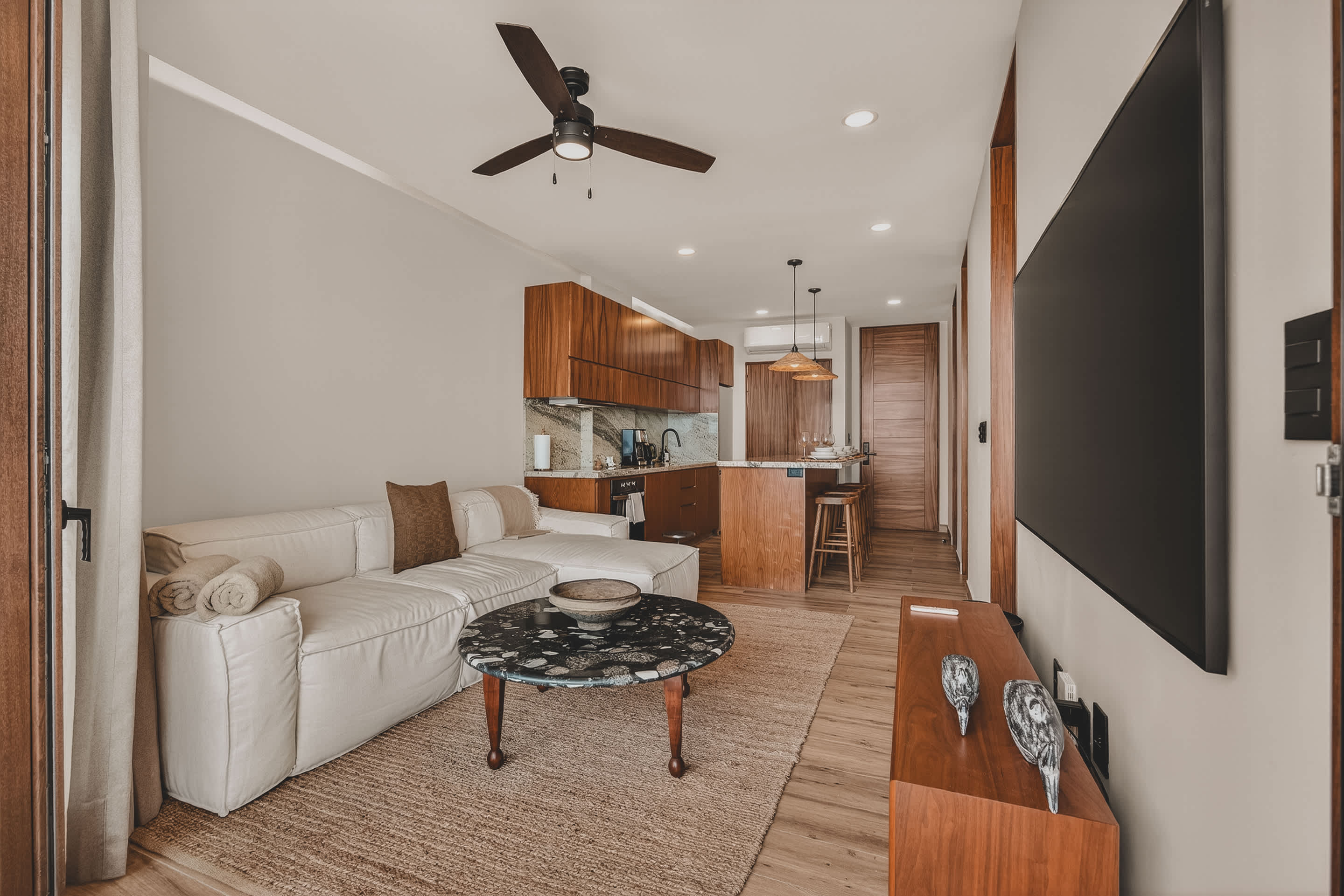 An open-concept living area with a large white sectional sofa, a dark circular coffee table, and a woven rug. In the background, a modern kitchen with wooden cabinetry, a breakfast bar with stools, and a ceiling fan are visible.