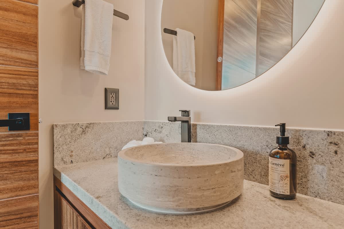 A close-up of a contemporary bathroom vanity featuring a light stone vessel sink, a black modern faucet, and a granite countertop. A large round backlit mirror is mounted above, and a bottle of soap sits on the counter.