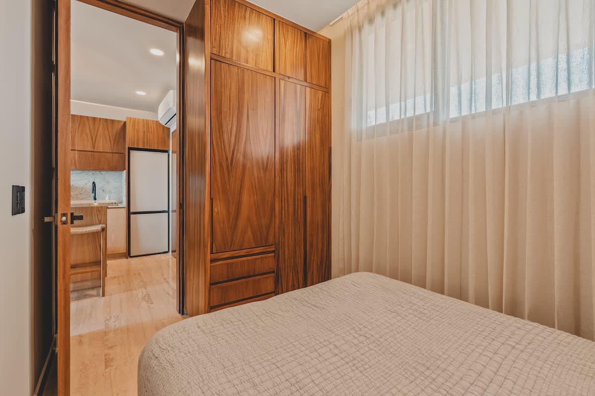 A view into a bedroom with a light-colored quilted bed, a large built-in wooden wardrobe, and sheer curtains covering a window. A doorway reveals a glimpse of a modern kitchen with a refrigerator and bar seating.