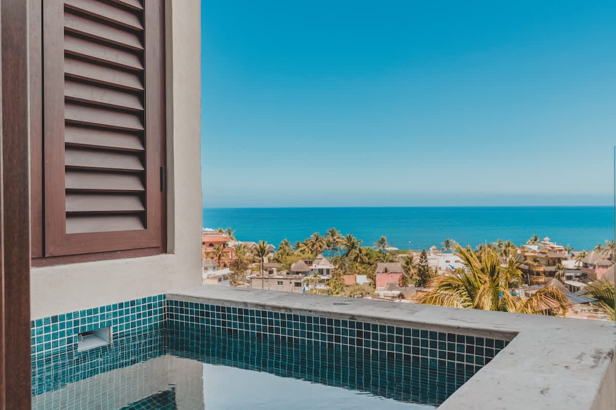A private balcony hot tub with blue mosaic tiles offers stunning panoramic ocean and town views under a clear blue sky, with a wooden louvered window visible on the left.