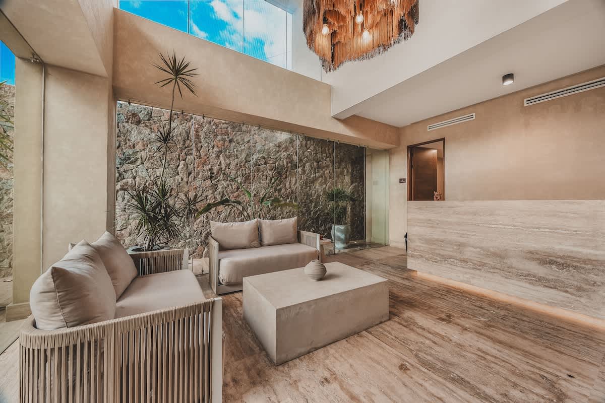 Lobby area featuring a stone accent wall, two light-colored sofas, a concrete coffee table, and large glass windows looking out to lush greenery. Two large, dark, textured chandeliers hang from the high ceiling.