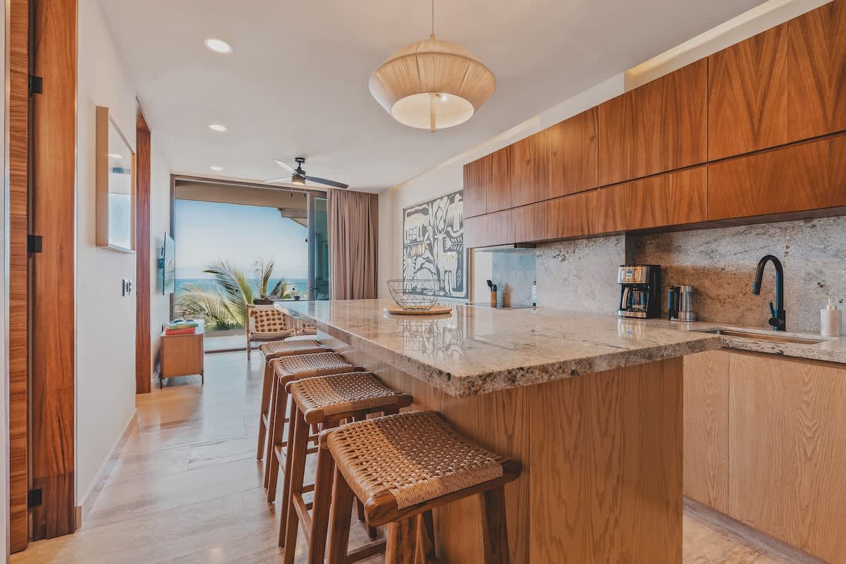Contemporary kitchen featuring a long granite-topped island with four woven bar stools, light wood cabinetry, and a stylish pendant light. Beyond the kitchen, a living area and ocean views are visible.