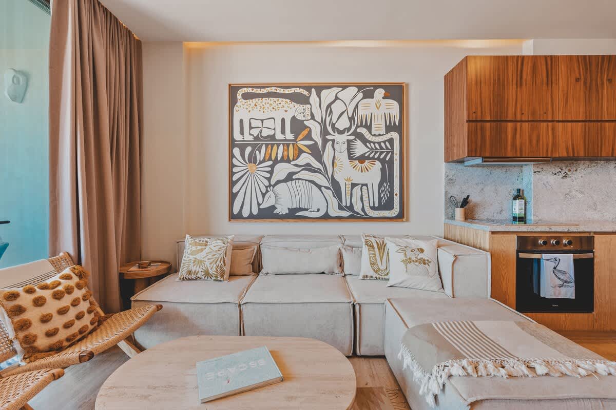 A chic living room features a large L-shaped sectional sofa with decorative pillows, a round light-colored coffee table, and a woven armchair with a patterned cushion. A large, colorful abstract artwork with animal motifs hangs above the sofa, and a modern kitchen area is visible in the background.