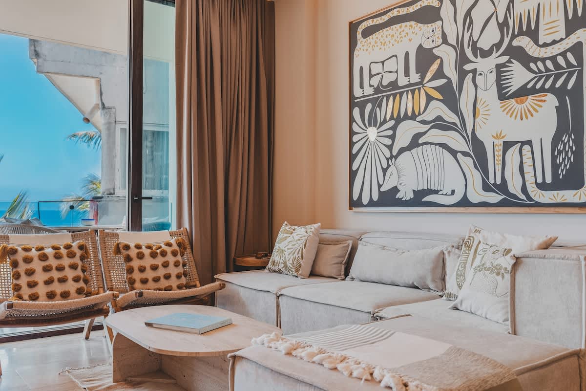 A chic living room features a large L-shaped sectional sofa with decorative pillows, a light wood coffee table, and two woven armchairs, all set against a large abstract artwork and offering ocean views through a glass door.