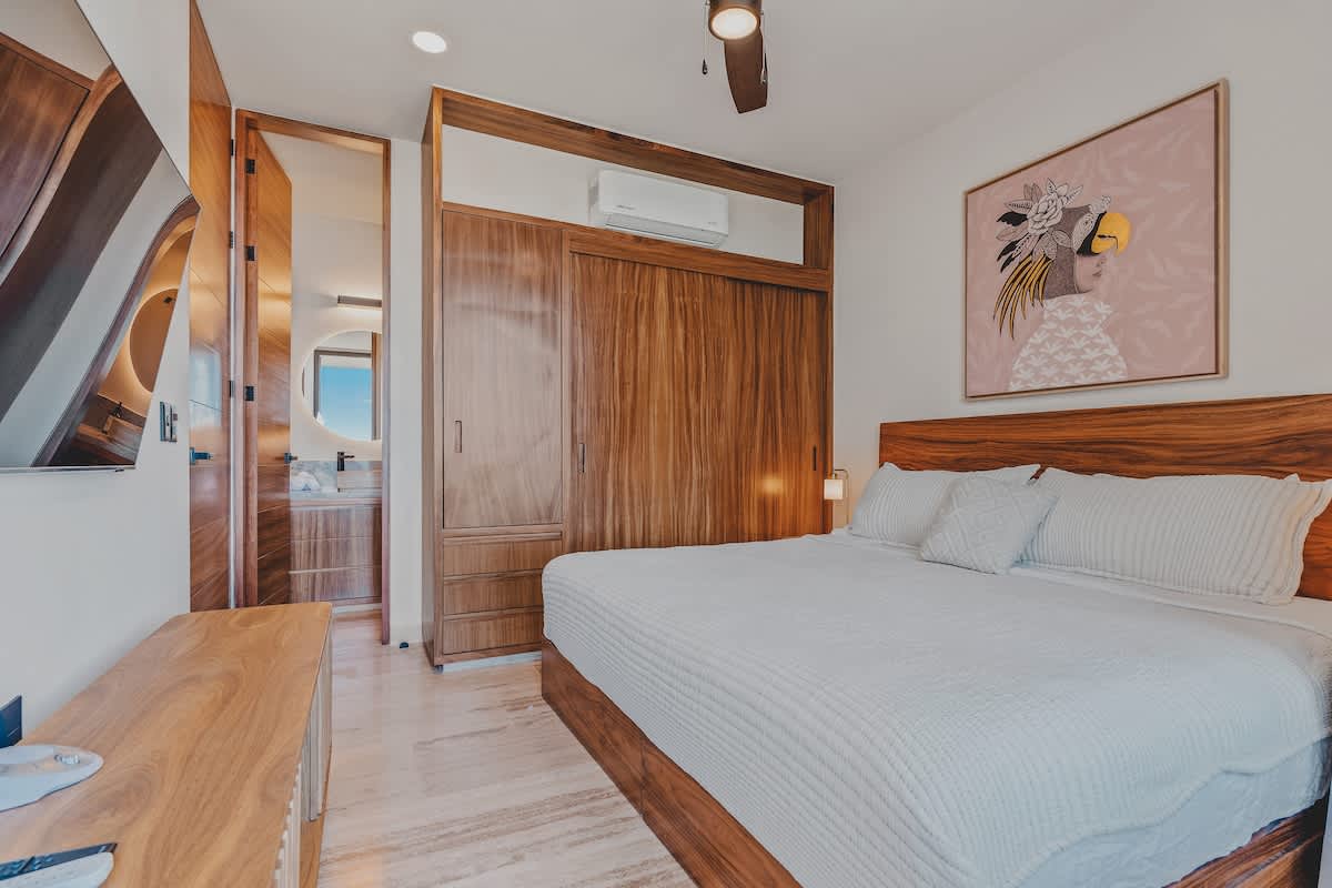 A bright bedroom features a king-sized bed with a wooden frame and headboard, and a large built-in wooden wardrobe with sliding doors. A wall-mounted television is visible on the left, and a doorway leads to an en-suite bathroom with a round backlit mirror.