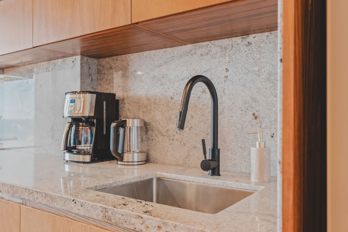 A modern kitchen countertop features light granite, a black gooseneck faucet over a stainless steel sink, and a coffee maker and kettle, with warm wooden cabinetry above.