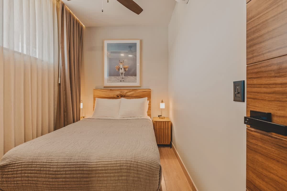 A cozy bedroom features a full-sized bed with a light brown quilted cover, a natural wood headboard, and matching bedside tables with lamps. A framed artwork of a donkey with flowers adorns the wall above the bed, and sheer and opaque curtains cover a large window.