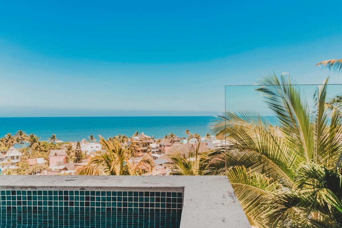 A close-up view of the balcony hot tub's blue mosaic tiles, showcasing a breathtaking panoramic ocean and town vista under a clear azure sky, with lush palm trees in the foreground.