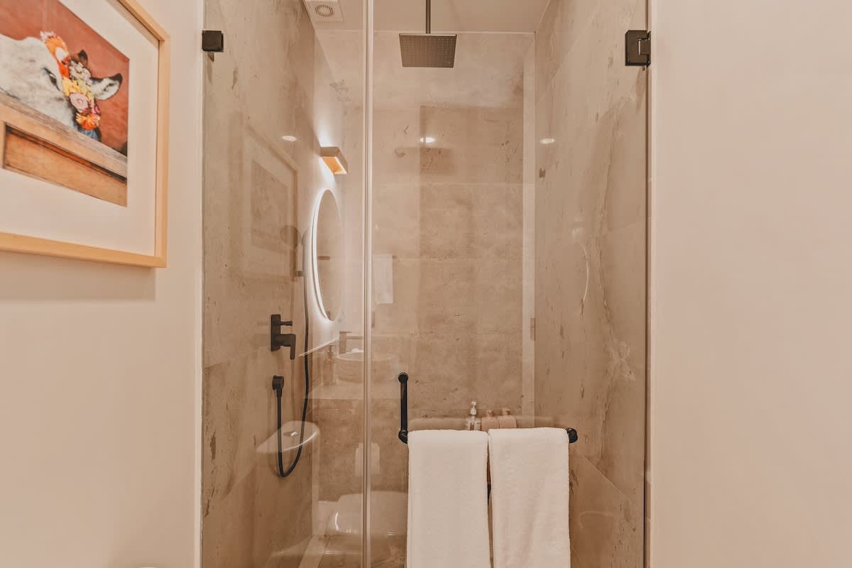 A sleek walk-in shower with clear glass doors and light-colored tiled walls features a rainfall showerhead and a handheld shower. Fresh white towels are neatly folded on a black towel bar inside the shower.