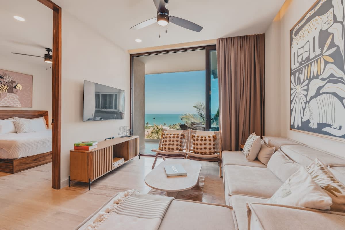 Modern living room with a light-colored sectional sofa, wooden coffee table, and a large abstract art piece. Sliding glass doors offer panoramic ocean views, connecting to a balcony.