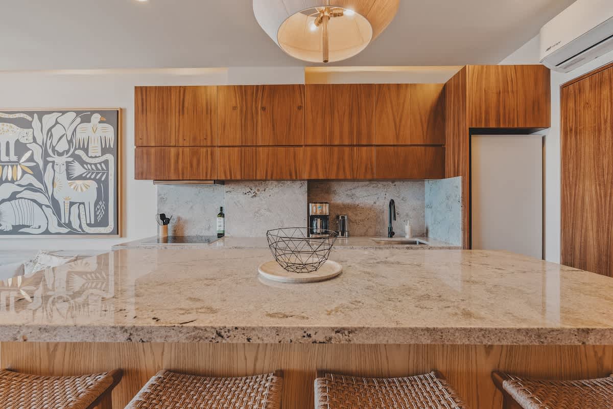 Detailed view of the kitchen island with a granite countertop and a decorative wire fruit bowl. The background shows dark wood upper cabinets, a light granite backsplash, and a large piece of abstract artwork.