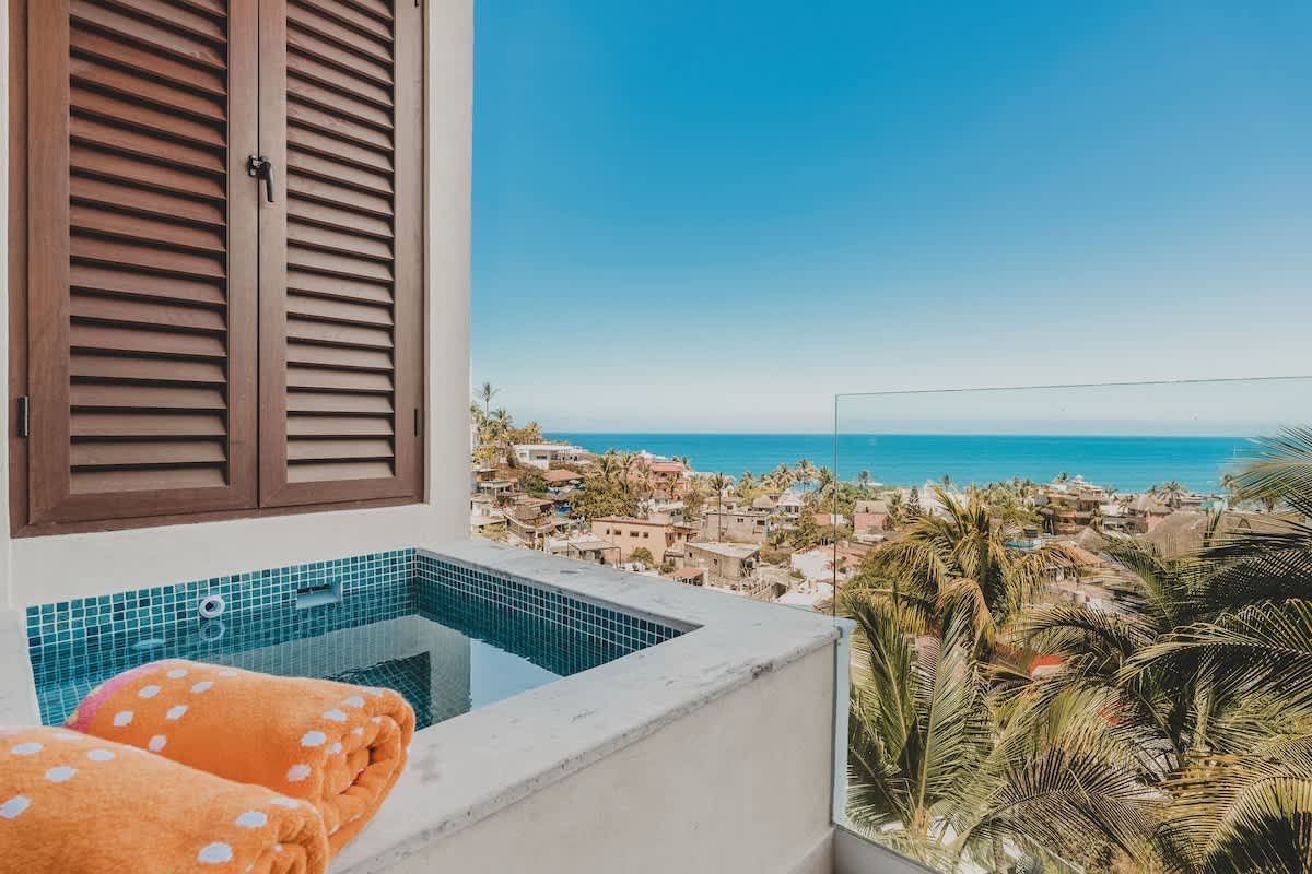 A private balcony hot tub with blue mosaic tiles and rolled orange towels offers expansive ocean and town views, framed by a wooden louvered window and a clear glass railing.