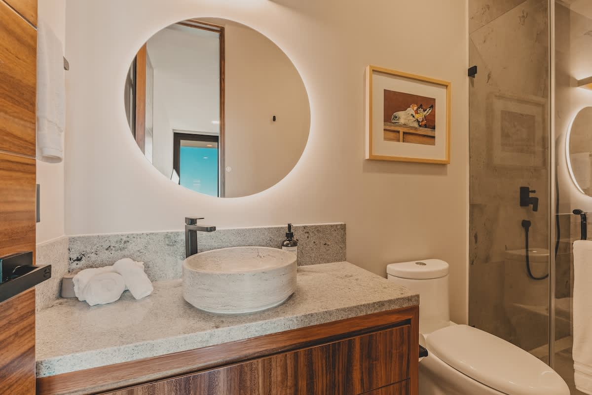 A modern bathroom features a light-colored stone vessel sink on a matching granite countertop, set against a dark wood vanity. A large round backlit mirror hangs above the sink, and a toilet is visible to the right.