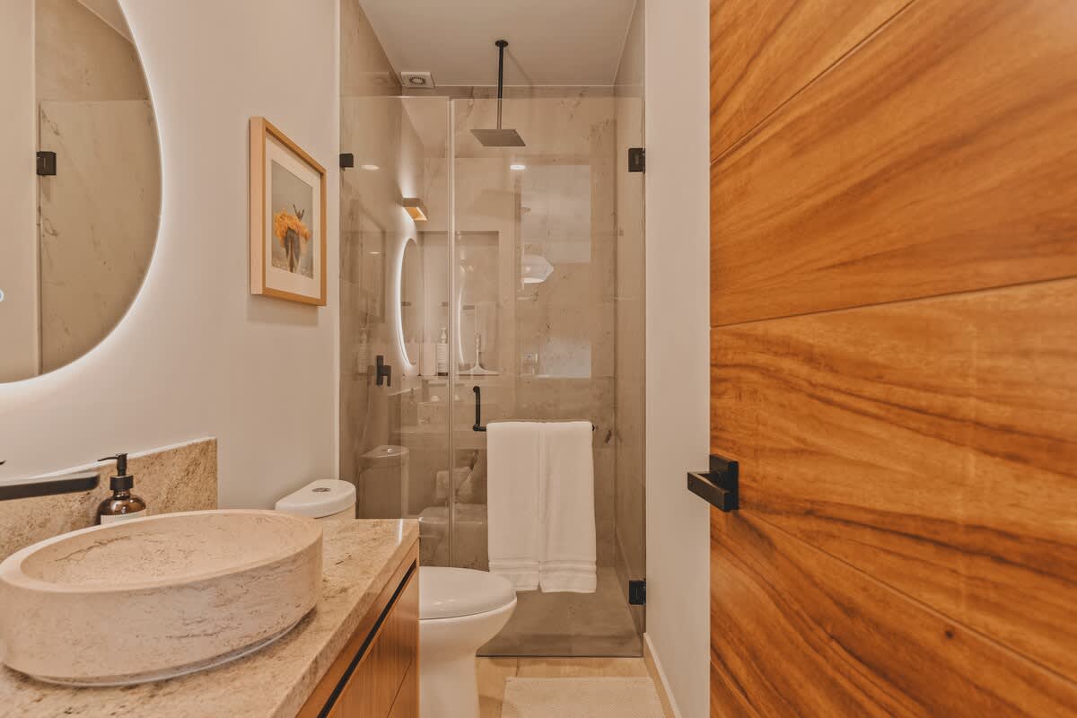 Contemporary bathroom featuring a glass-enclosed walk-in shower with a rainfall showerhead and a modern toilet. The vanity has a stone vessel sink and a large backlit circular mirror.