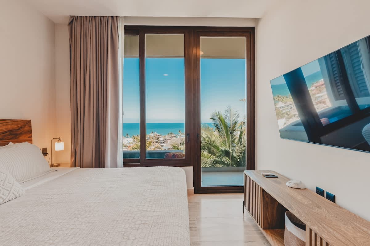 A bright bedroom featuring a large bed with a natural wood headboard, light bedding, and a wall-mounted TV. Floor-to-ceiling glass doors offer stunning ocean views with palm trees and distant buildings.