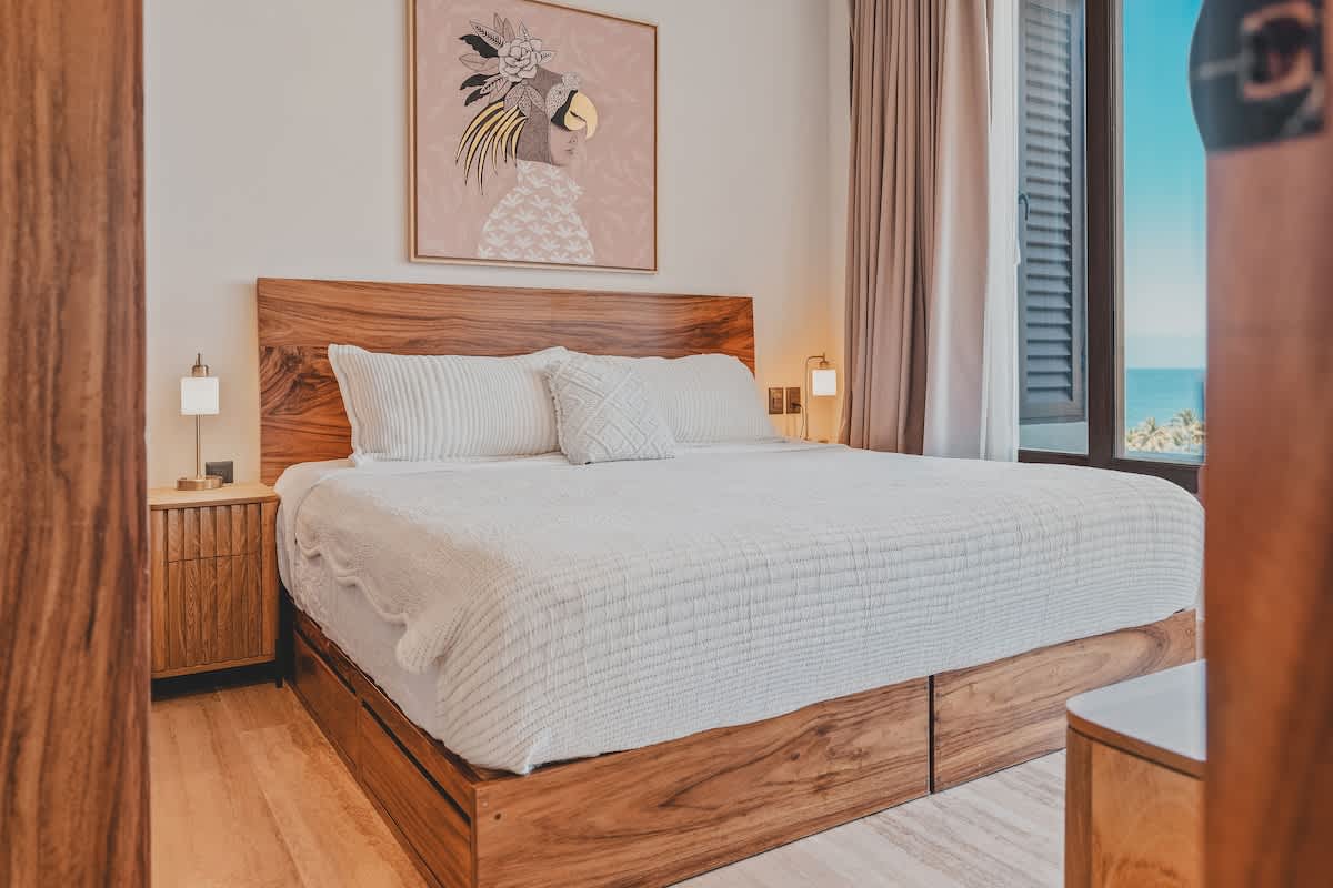 A spacious bedroom features a king-sized bed with a natural wood frame and headboard, dressed in white bedding. Modern bedside tables with lamps flank the bed, and a large framed artwork with a stylized figure hangs above the headboard. Ocean views are visible through the window.
