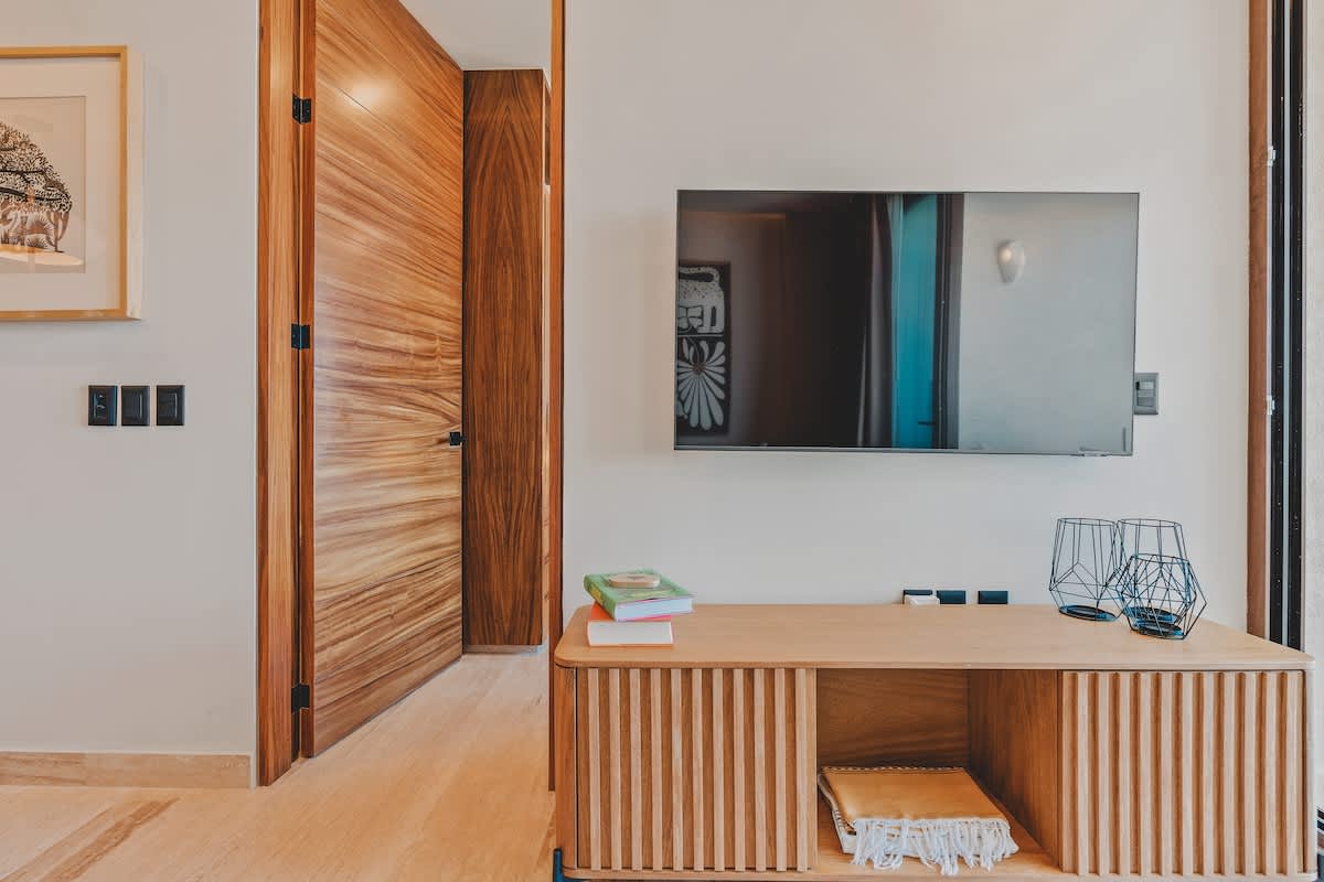 A modern living room wall features a wall-mounted flat-screen TV above a slatted wooden media console, with a warm wooden door leading to another room and minimalist decor.