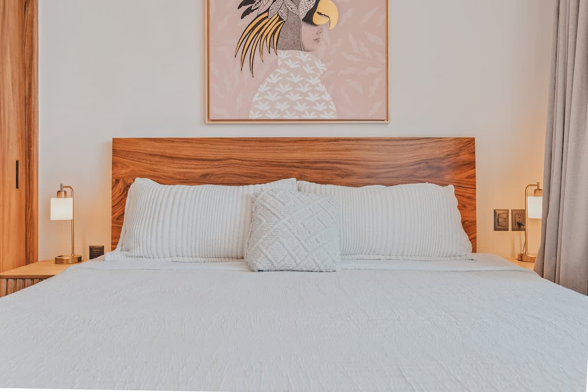 A close-up view of a king-sized bed with a natural wood headboard and white quilted bedding, accented by a decorative throw pillow. A vibrant framed artwork depicting a stylized figure with a bird-like headdress hangs above the bed.