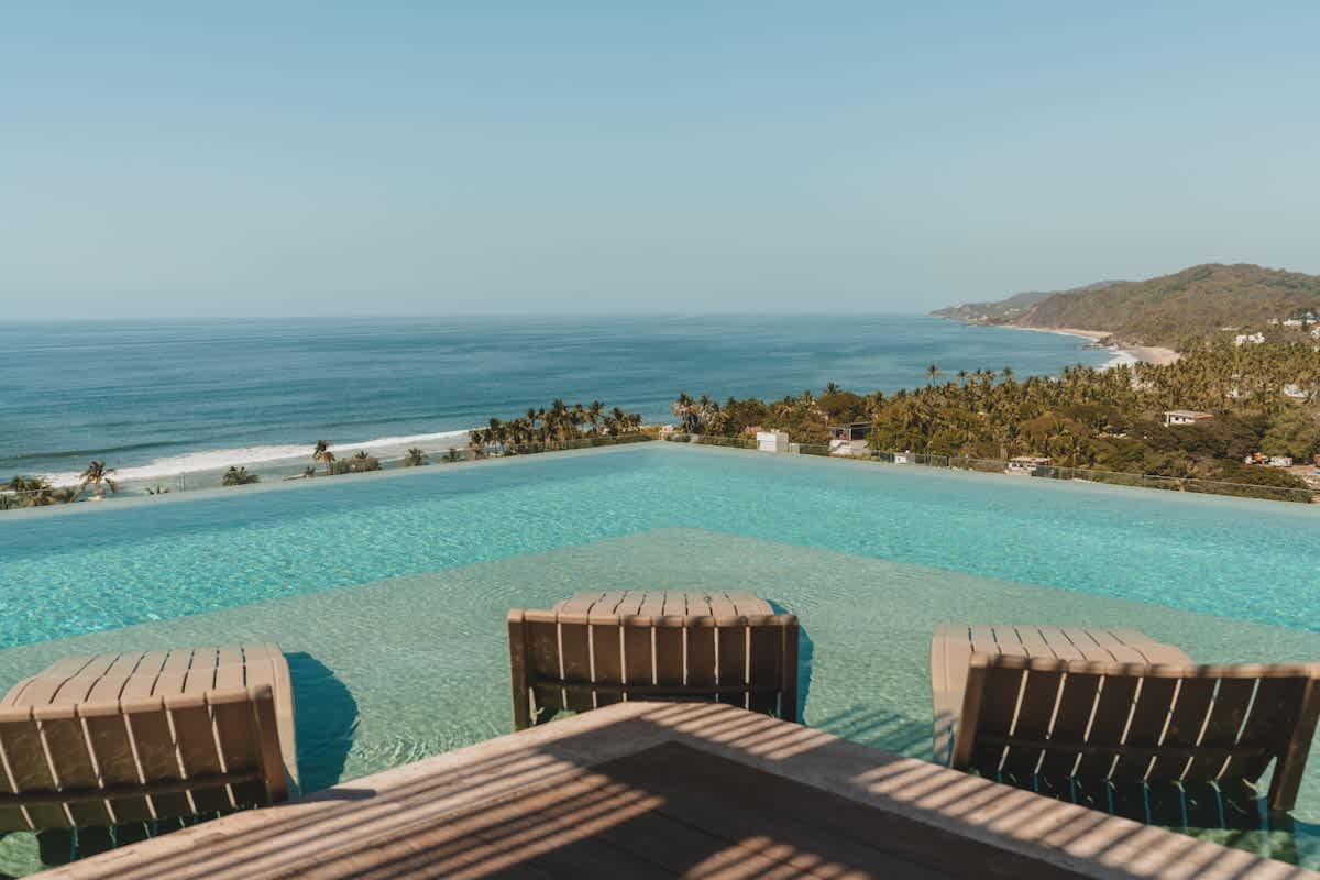 A stunning infinity pool stretches towards the horizon, featuring submerged lounge chairs at its edge, offering an unobstructed, expansive view of the deep blue ocean and a distant green coastline.