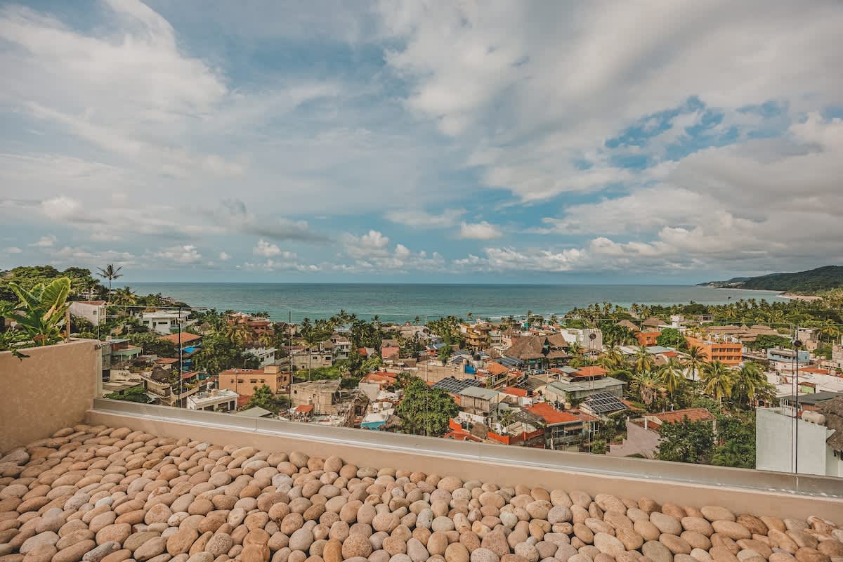 An expansive rooftop deck with light-colored river rocks and a low wall provides breathtaking views of the ocean meeting a coastal town with lush greenery under a bright, cloudy sky.