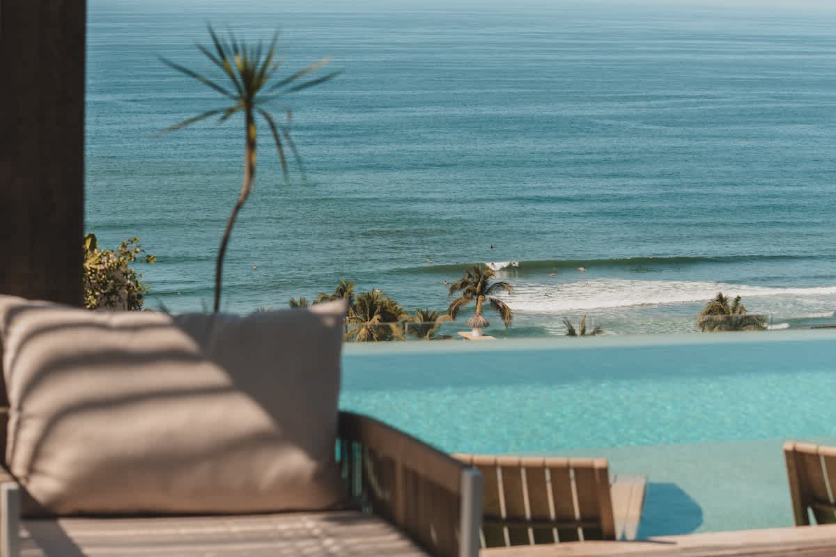 A comfortable outdoor lounge chair with a beige cushion sits beside an infinity pool, overlooking the vast blue ocean with distant waves and lush palm trees under a clear sky.