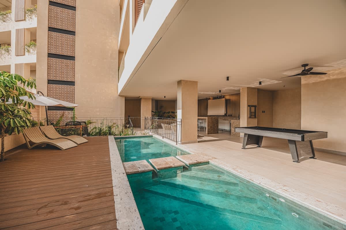 An outdoor common area features a long, narrow swimming pool with stepping stones, flanked by a wooden deck with two lounge chairs and an umbrella. In the background, the same grey pool table is visible under a covered patio with a ceiling fan and light-colored walls.