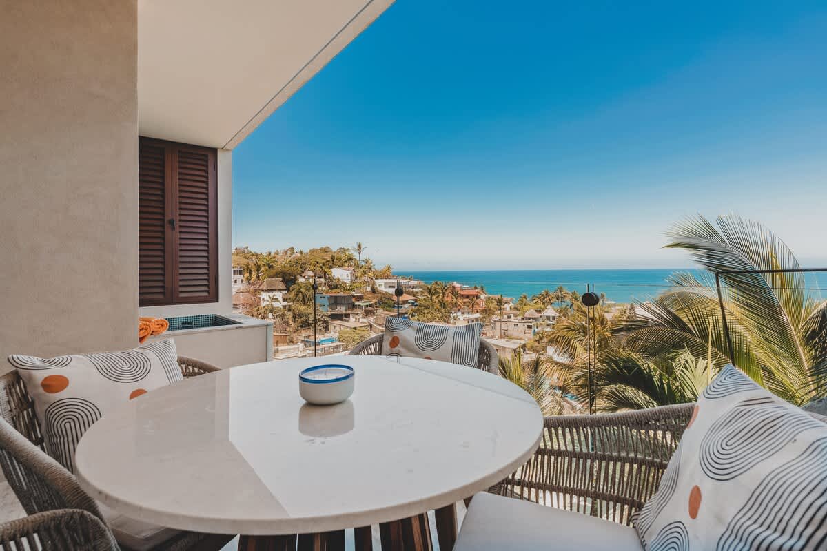 Expansive balcony with a round outdoor dining table and chairs, providing stunning panoramic views of the vibrant coastal town, lush palm trees, and the sparkling ocean. A private hot tub is visible to the side.