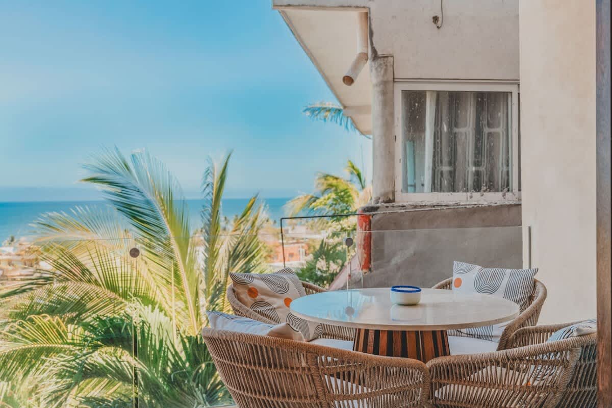A charming outdoor dining area on a balcony features a round marble-top table and four woven chairs with patterned cushions, offering a partial ocean view framed by lush palm trees.