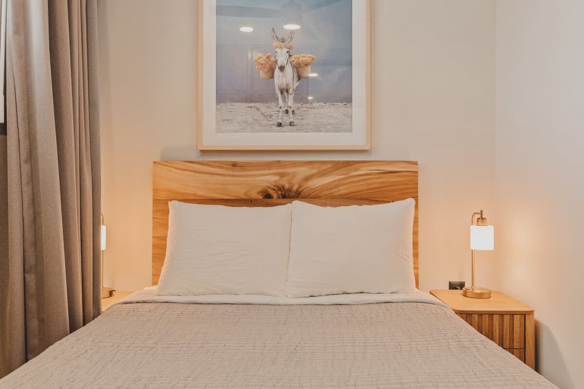 Detailed view of a comfortable queen bed with a unique natural wood headboard and crisp white pillows. Two modern table lamps flank the bed, and a whimsical artwork is displayed above.