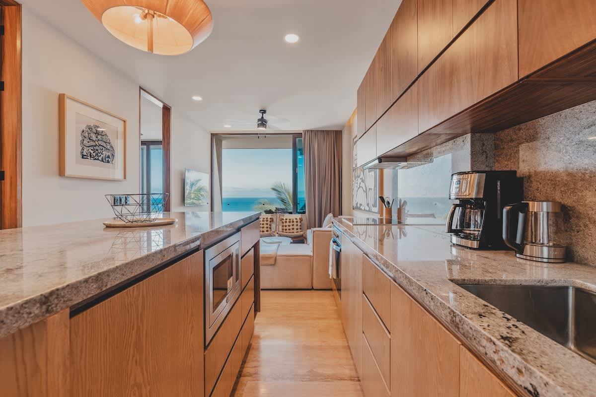 Modern kitchen with light wood cabinetry, granite countertops, and stainless steel appliances, including a built-in microwave. The space opens up to a living area with ocean views visible through large glass doors.