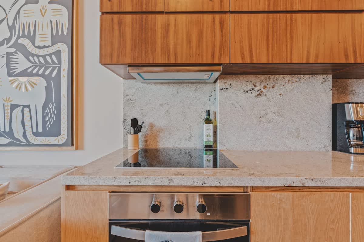 Close-up of the kitchen counter featuring a sleek black electric cooktop, a stainless steel built-in oven, and a light granite backsplash. An olive oil bottle and cooking utensils are neatly arranged.