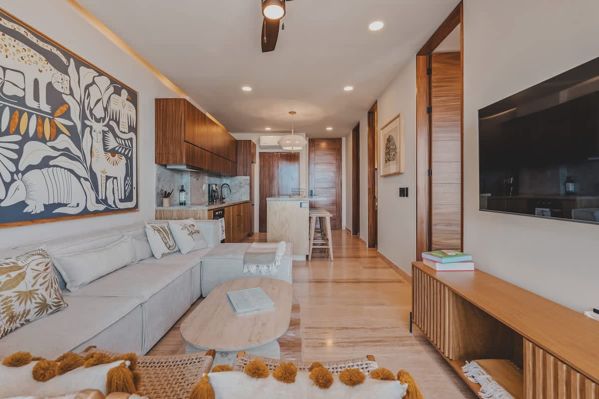 An open-concept living space features a light-colored sectional sofa, a round coffee table, and a wooden TV console with a mounted television. The adjacent modern kitchen boasts dark wood cabinetry, light countertops, and a breakfast bar with stools.