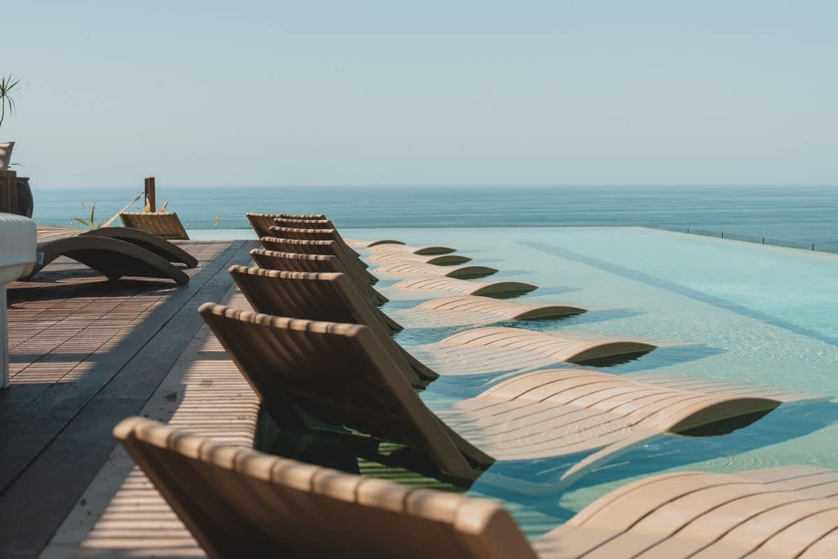 Serene close-up of an infinity pool with several wooden lounge chairs partially submerged in the clear blue water, seamlessly blending with the vast ocean horizon.
