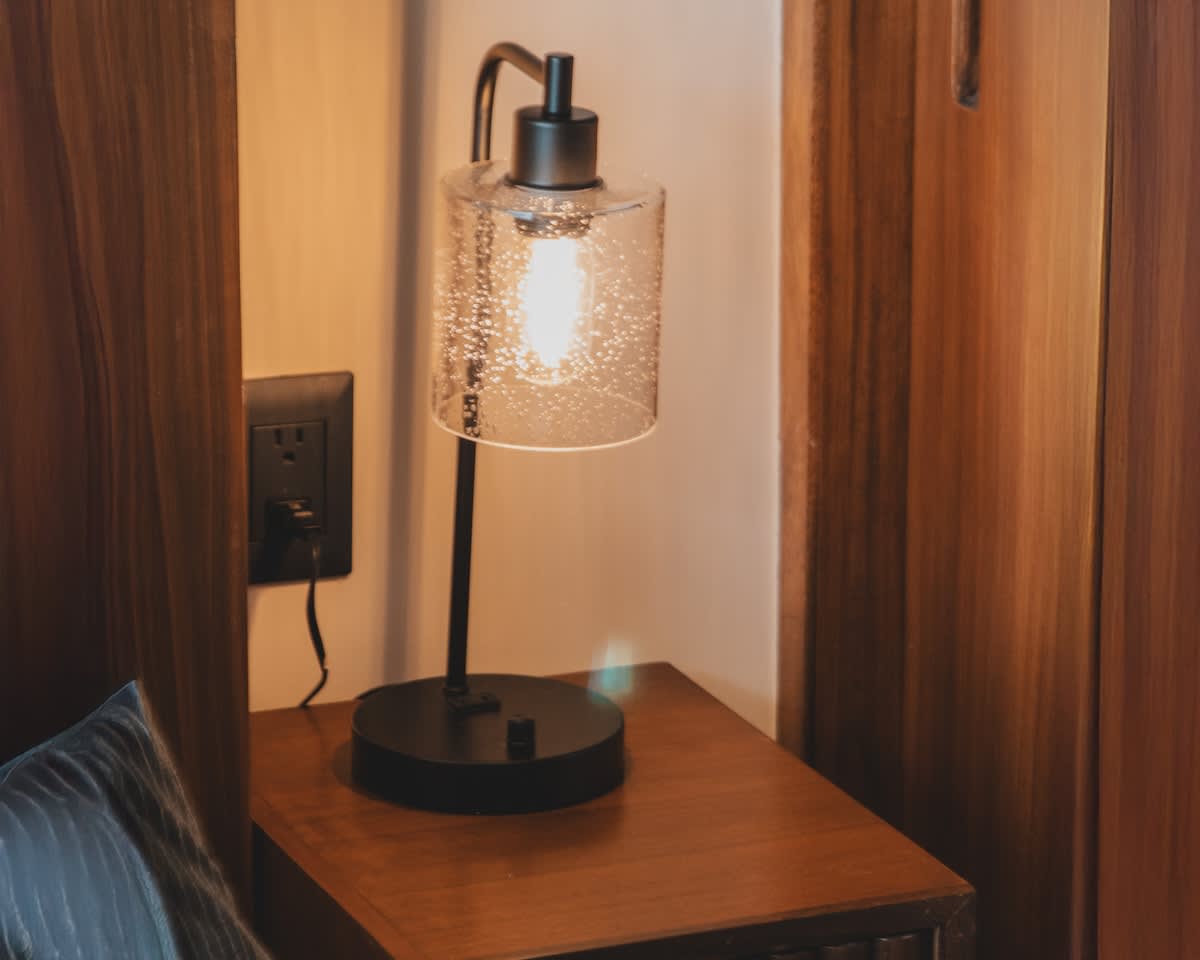 A modern black table lamp with a clear, seeded glass shade illuminates a dark wooden bedside table, positioned next to a power outlet in a cozy bedroom setting.