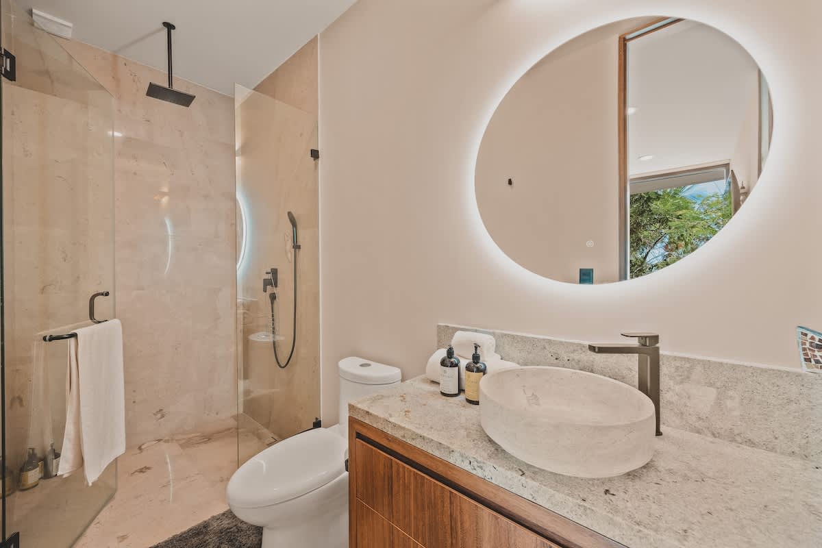 Modern bathroom featuring a glass-enclosed shower with a rainfall showerhead, a light-colored stone vanity with a vessel sink, and a large circular backlit mirror.