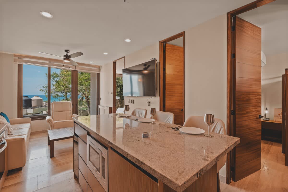 Bright living space with a large granite kitchen island, a light-colored sofa, and a wall-mounted TV, featuring a ceiling fan and expansive windows offering views of the tropical surroundings.