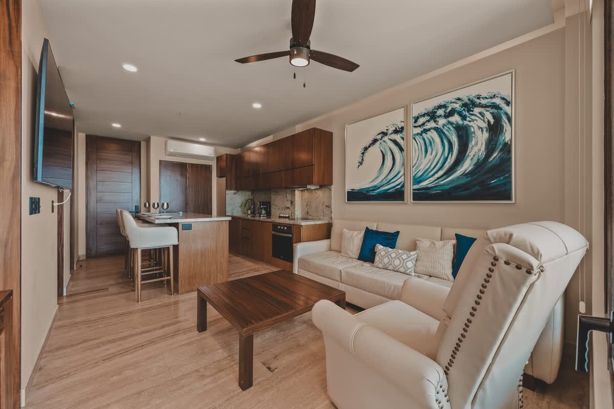 Open-concept living area with a white sofa, a recliner, a wooden coffee table, and two large wave-themed artworks, adjacent to a modern kitchen with dark wood cabinetry and a breakfast bar.