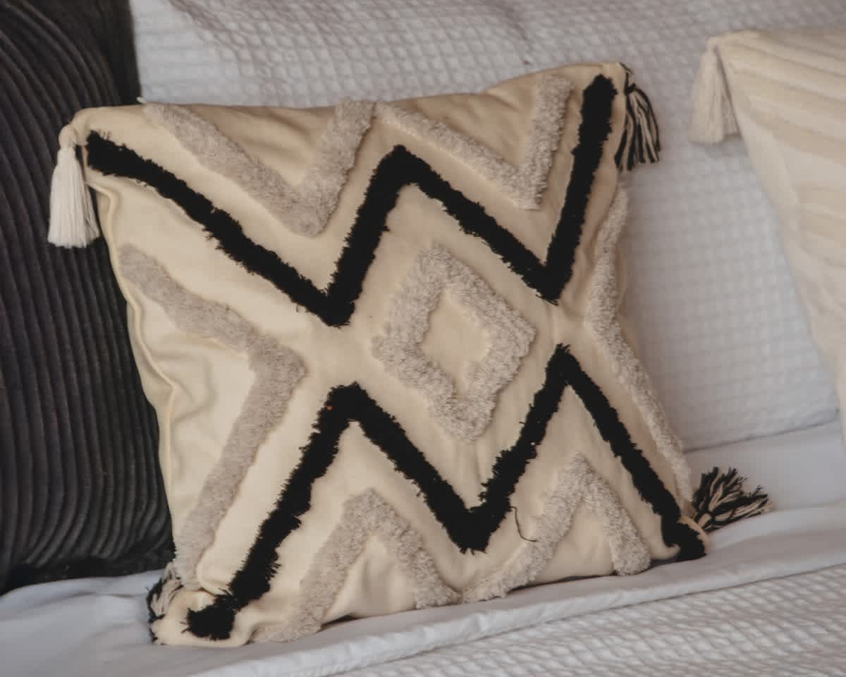 A decorative throw pillow with a cream background and bold black and textured beige zigzag and diamond patterns, accented with tassels, rests on a white bedspread, adding a touch of bohemian style.