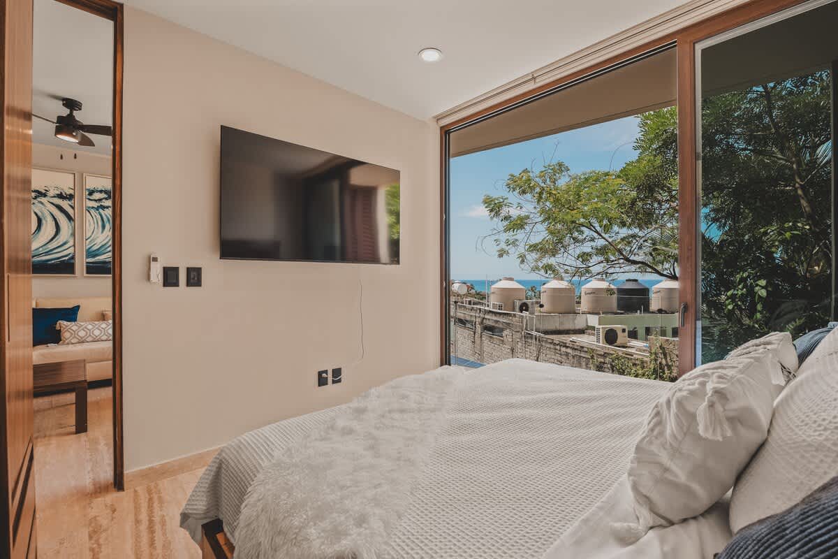 Modern bedroom with a large wall-mounted TV, a bed with white and grey bedding, and a wide sliding glass door providing expansive views of the tropical landscape and distant ocean.