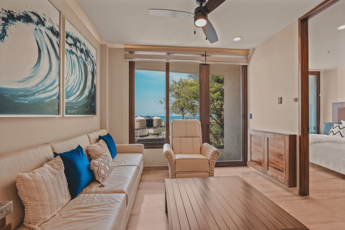 Bright living room with a white sofa, a recliner, and a wooden coffee table, featuring large windows offering a view of the ocean and a partial glimpse into an adjacent bedroom.