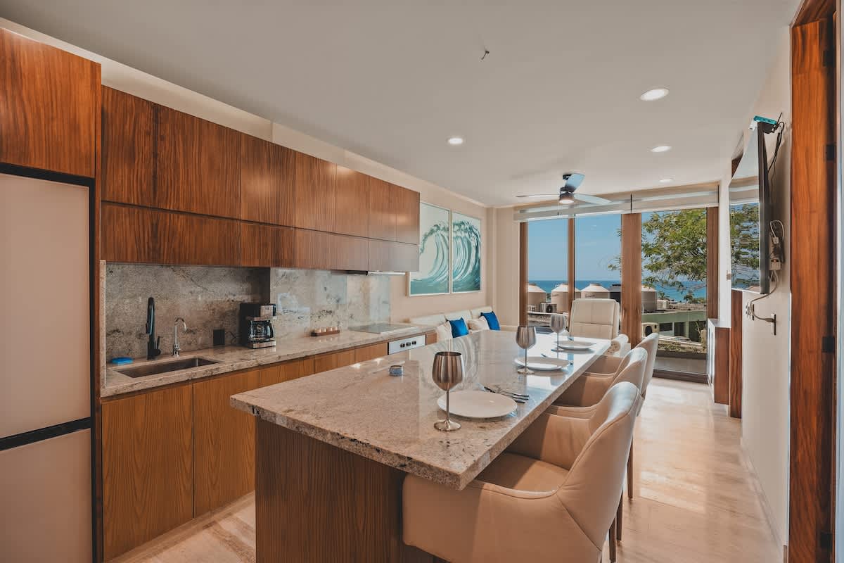Open-concept kitchen and dining area with a large granite island, white bar stools, and dark wood cabinetry, offering a glimpse of the living room and ocean views through a large window.