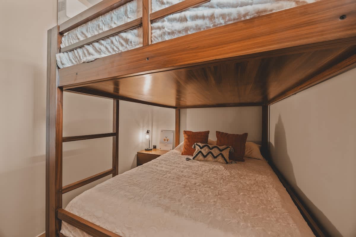 Detailed view of the lower bunk bed, showcasing a comfortable white bedspread, decorative pillows, a small wooden nightstand with a lamp, and the sturdy wooden frame of the upper bunk.