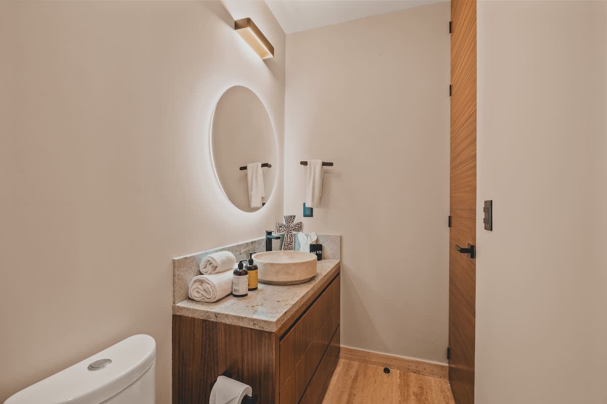 Contemporary bathroom with a light-colored stone vanity, a round vessel sink, a backlit circular mirror, and a toilet, all set against light-colored walls.