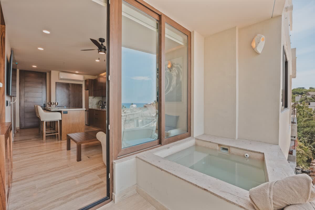 Balcony featuring a built-in hot tub with towels, offering a partial view of the ocean and the surrounding town, accessible from the living area through a large sliding glass door.