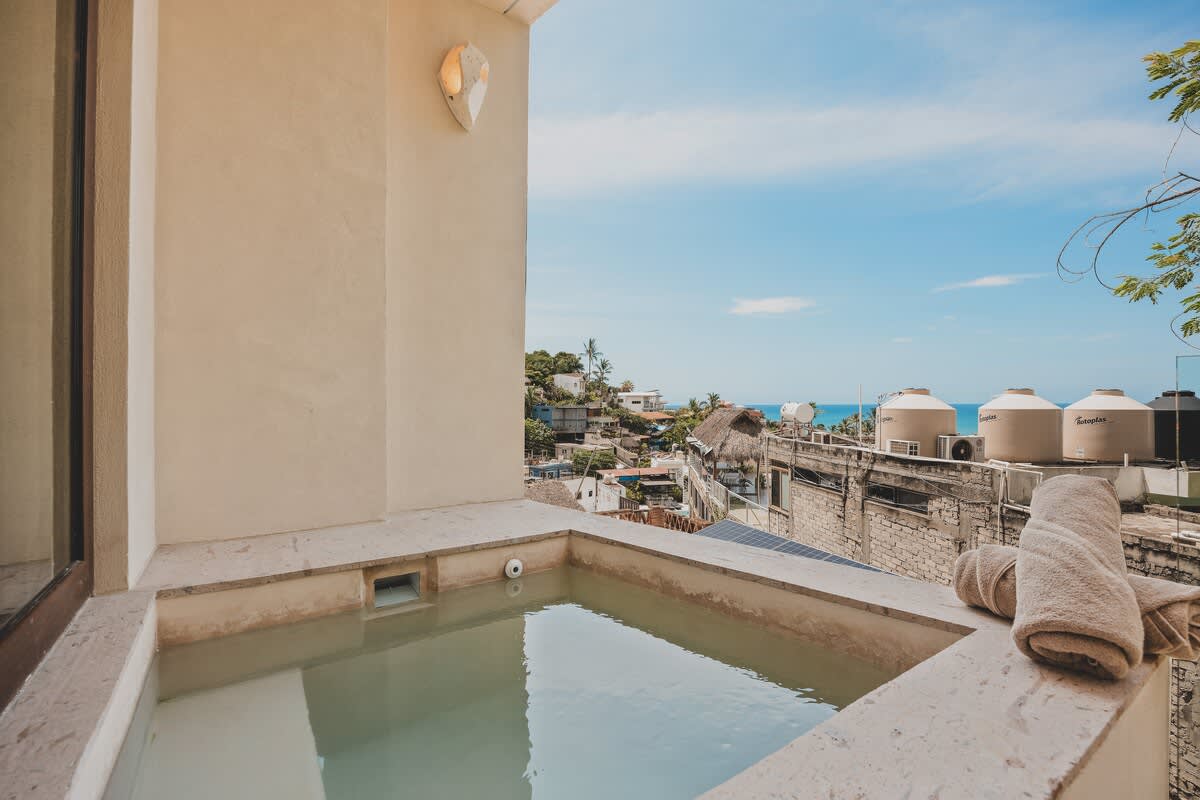 Outdoor balcony with a built-in hot tub, offering a scenic view of the coastal town's rooftops and the ocean under a bright blue sky, with towels neatly placed beside the tub.