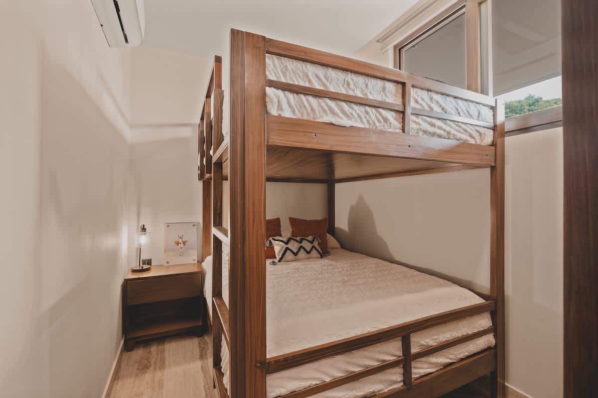 A cozy bedroom featuring a sturdy wooden bunk bed with white bedding, a small wooden nightstand with a lamp, and light-colored walls, creating a simple and functional sleeping space.