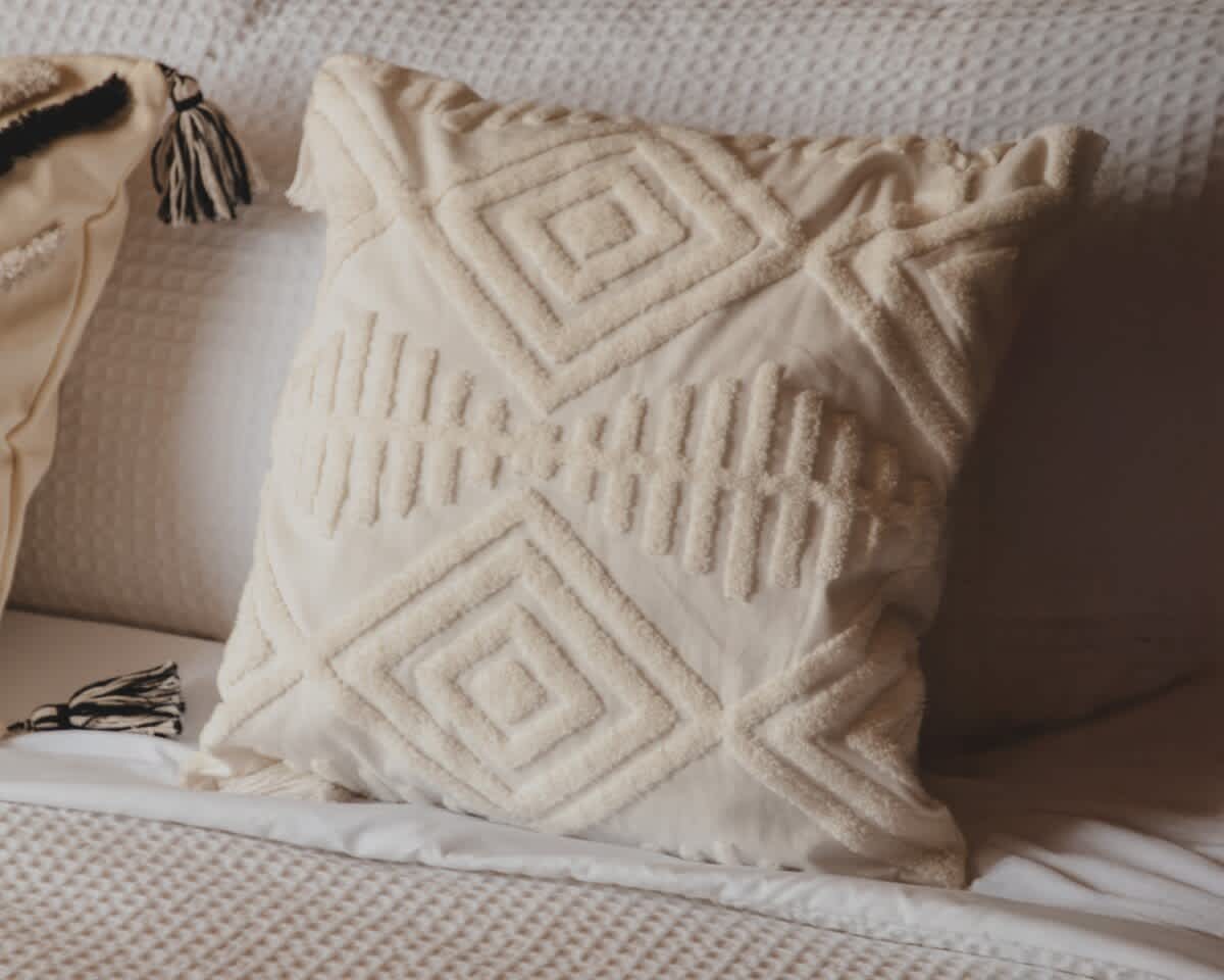 Cream-colored decorative pillow with raised geometric patterns, including diamonds and vertical lines, resting on a white textured bedspread, suggesting a cozy bedroom aesthetic.