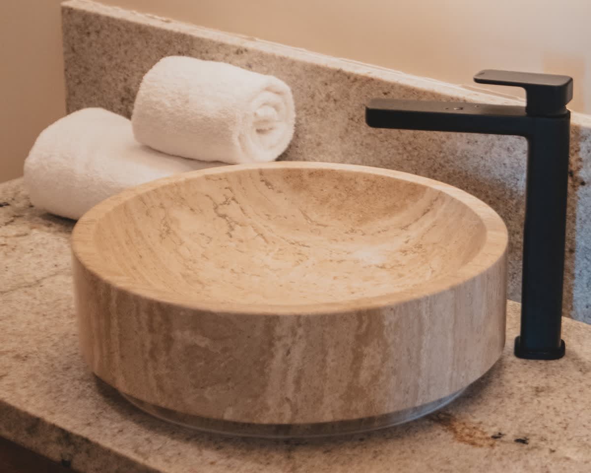 A round, light-colored stone vessel sink with natural veining sits on a granite countertop, paired with a sleek black faucet and two rolled white towels in a contemporary bathroom.