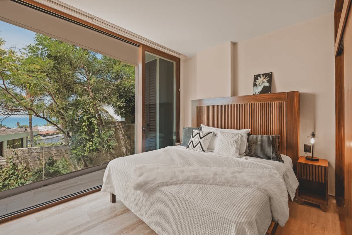 Master bedroom with a queen-sized bed featuring a slatted wooden headboard and white bedding, complemented by a wooden nightstand and a large sliding glass door opening to lush greenery.